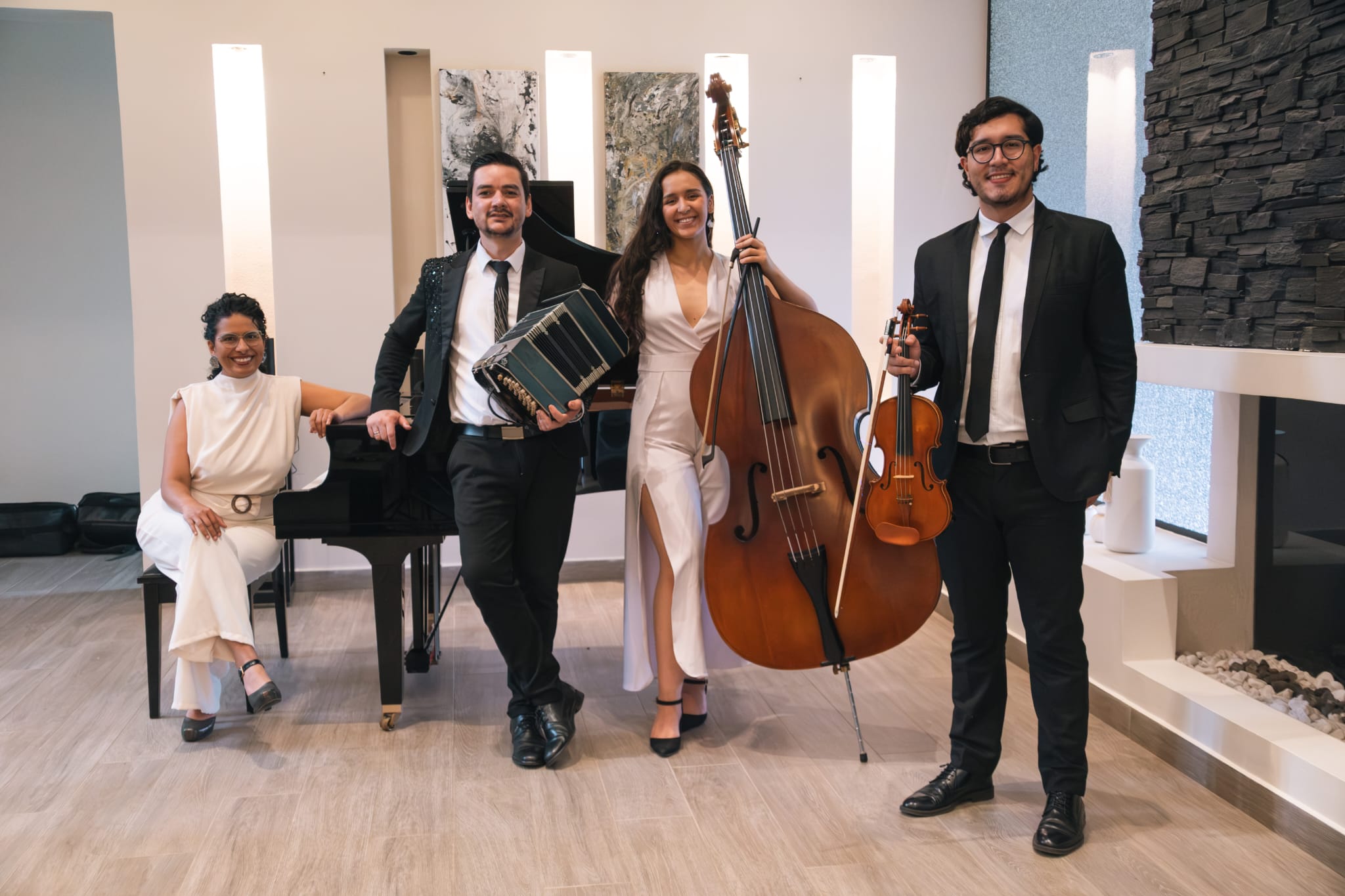 Four musicians in formal dress pose with piano, accordion, cello, and violin in a modern recital room.