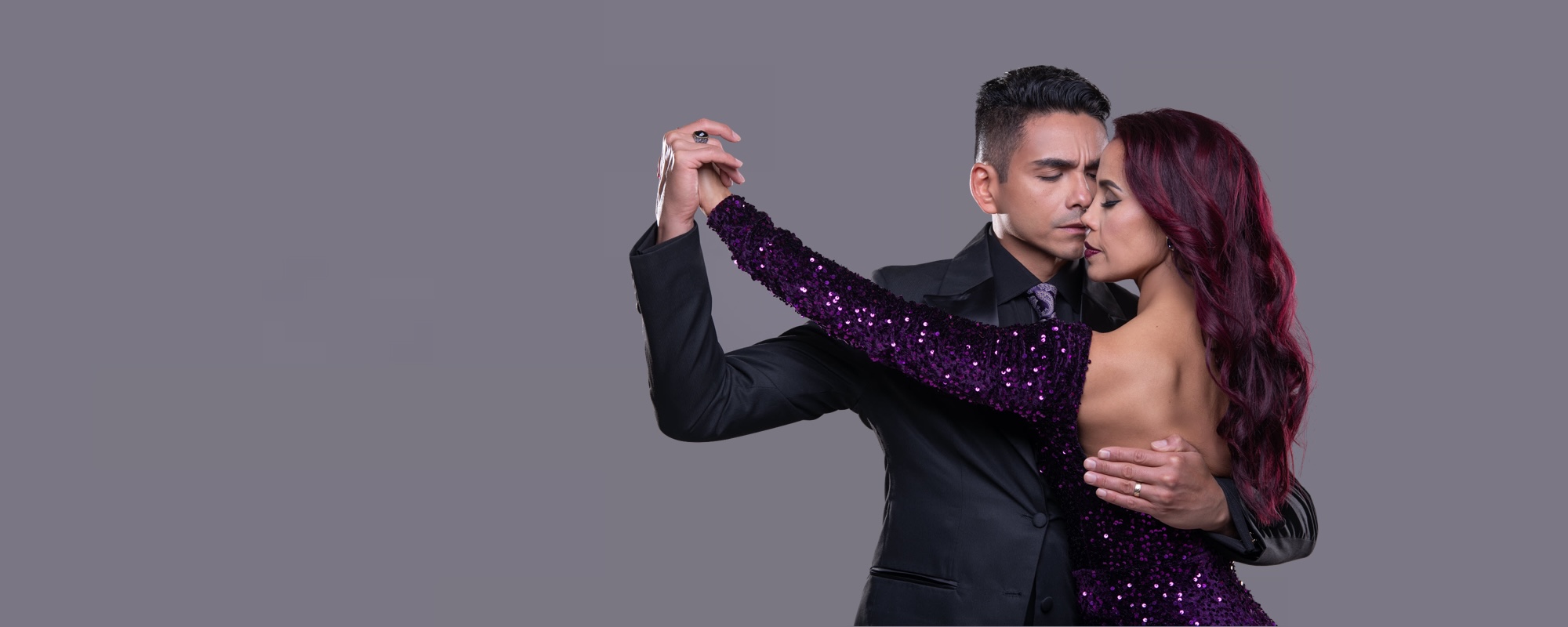 Tanzend logo tanya20 | Blanco y Negro Tango Couple dancing closely; woman in a purple sequined dress and man in a black suit, embracing with eyes closed against a gray background.