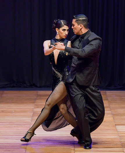 Couple dancing a ballroom routine on a wooden stage; woman in a black jeweled dress, man in a black suit, mid-step.