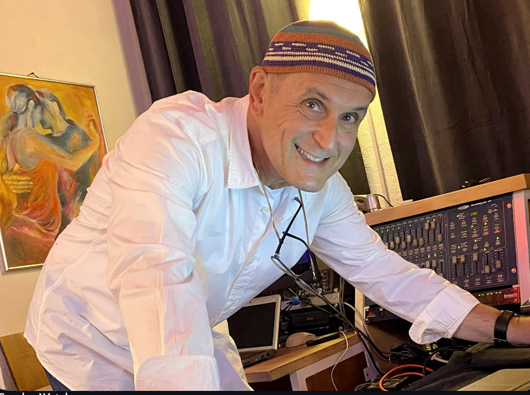 Smiling man in a white shirt and patterned cap leaning over an audio mixer in a recording studio.