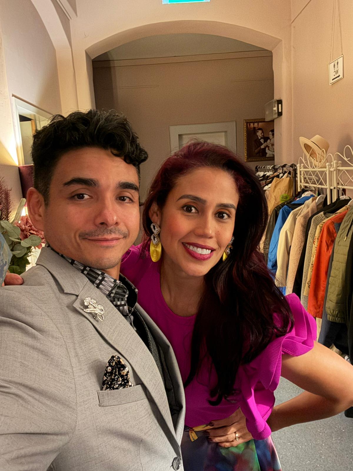 Two smiling adults pose for a selfie in a boutique, with racks of colorful clothes in the background.