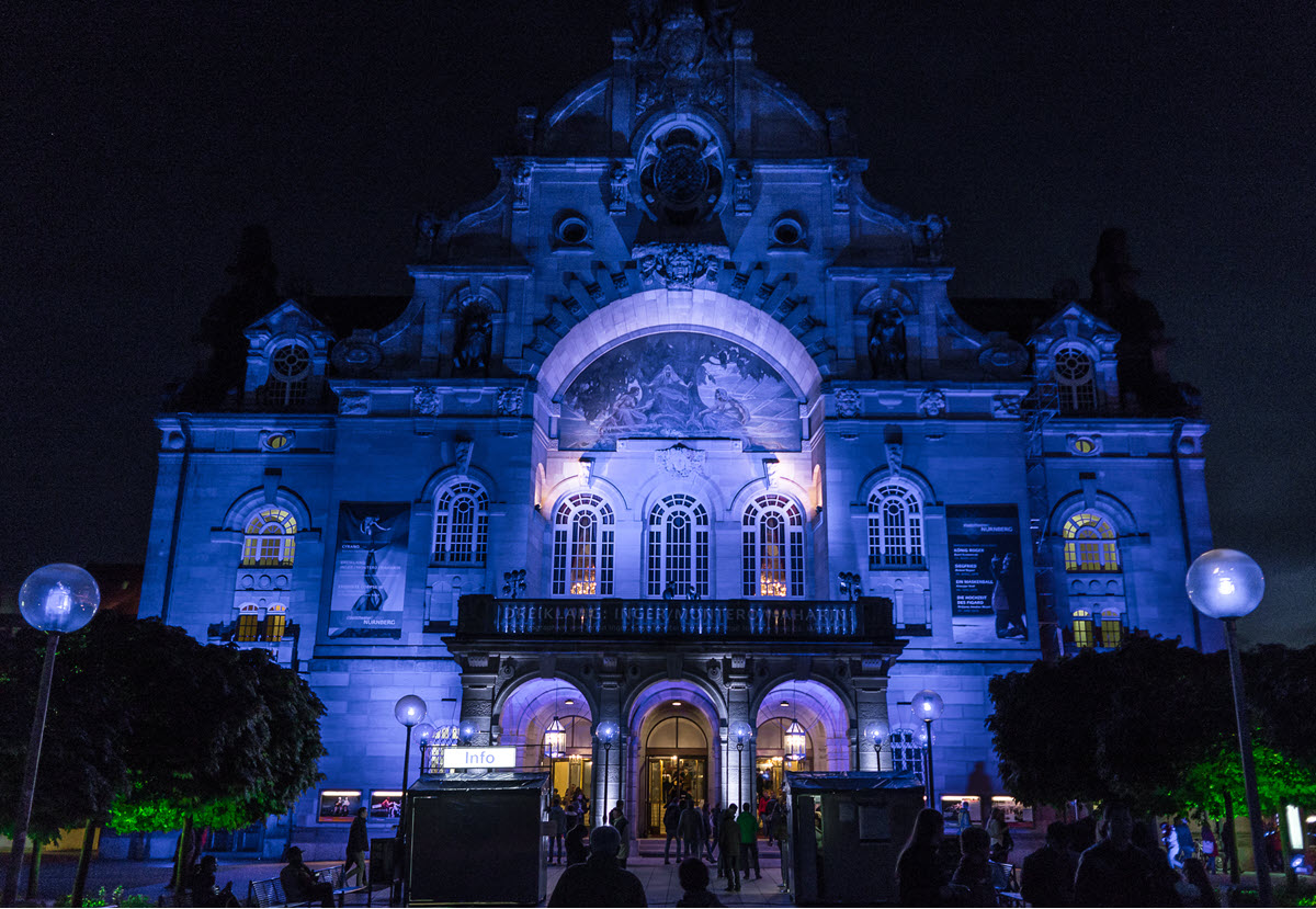 Night view of a grand historic building lit in blue, with arched entrances and people entering the doorway at dusk/dusk.