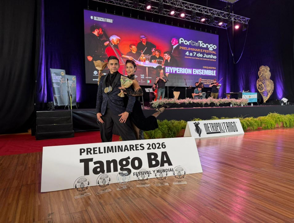 Tango couple in formal attire posing on a competition stage with a large banner reading PRELIMINARES 2026 Tango BA in the foreground.