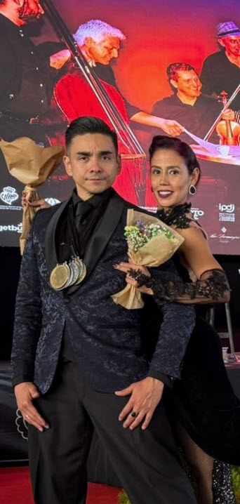 Couple posing on a red carpet with bouquets and medals, against a live music backdrop, celebrating an award achievement.
