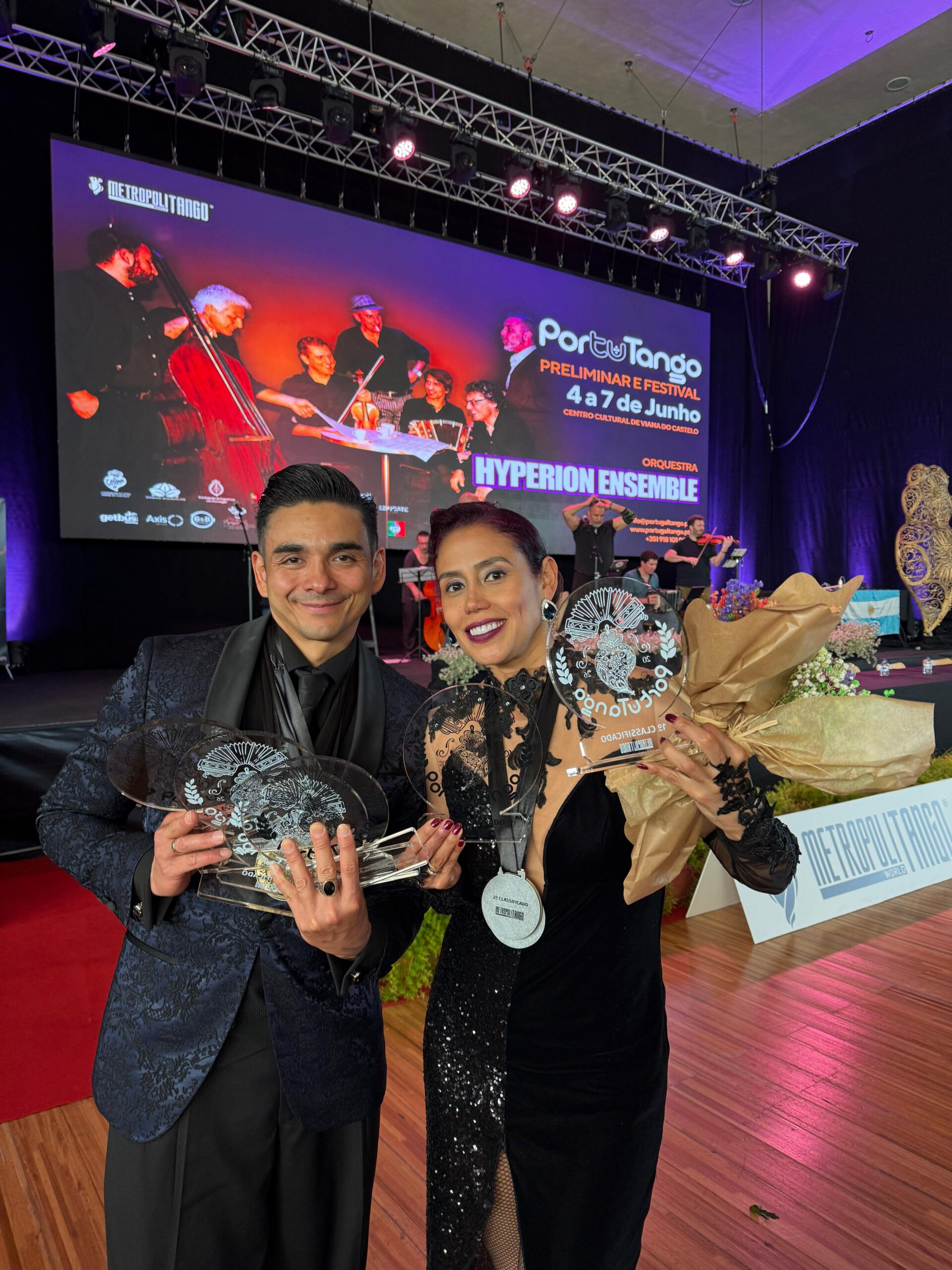 Two smiling performers in formal black outfits hold glass awards on a stage with a colorful festival backdrop behind them, Metropolitango banner visible.