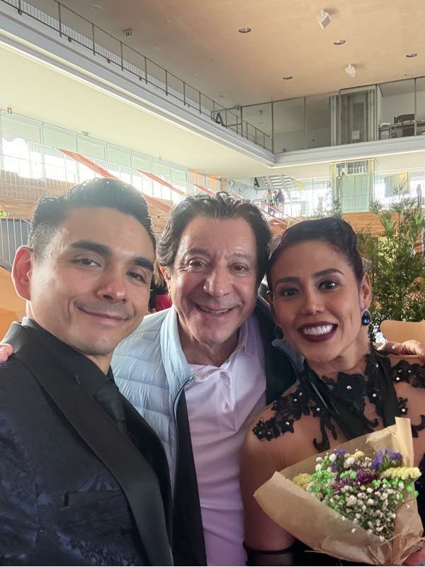 Three smiling people posing for a selfie in a bright indoor venue, with a bouquet held by the woman on the right.