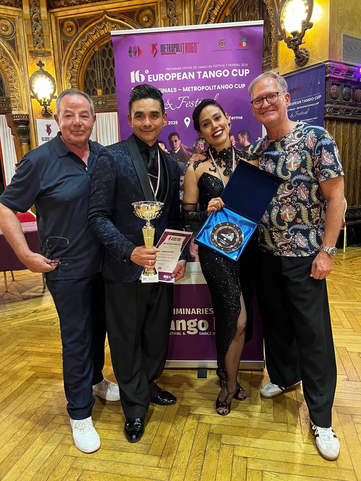 Claudio und Jörg sind dabei | Blanco y Negro Tango Four people pose with awards at the 16th European Tango Cup, in a decoratively ornate venue with a purple event banner behind them.