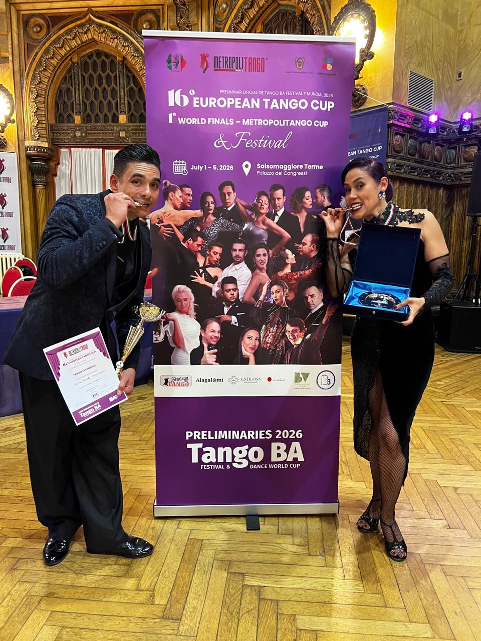 Siegerehrung 01 | Blanco y Negro Tango Two dancers pose with awards in front of a purple Tango event banner, holding a trophy and a certificate in a grand ballroom setting.