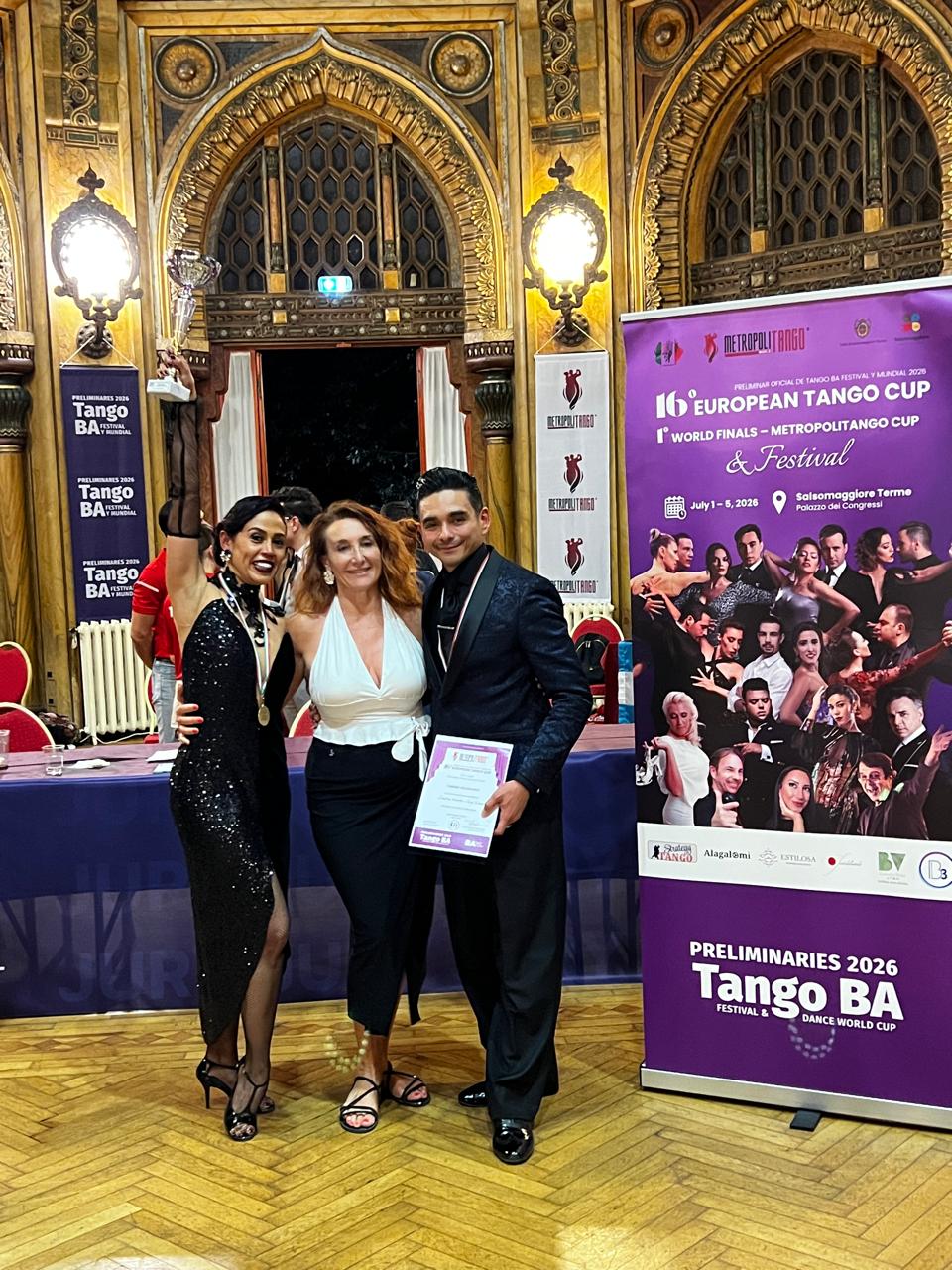 Siegerehrung 02 | Blanco y Negro Tango Three dancers pose with trophy and certificate in a gold ornate ballroom; banners show European Tango Cup and Tango BA.