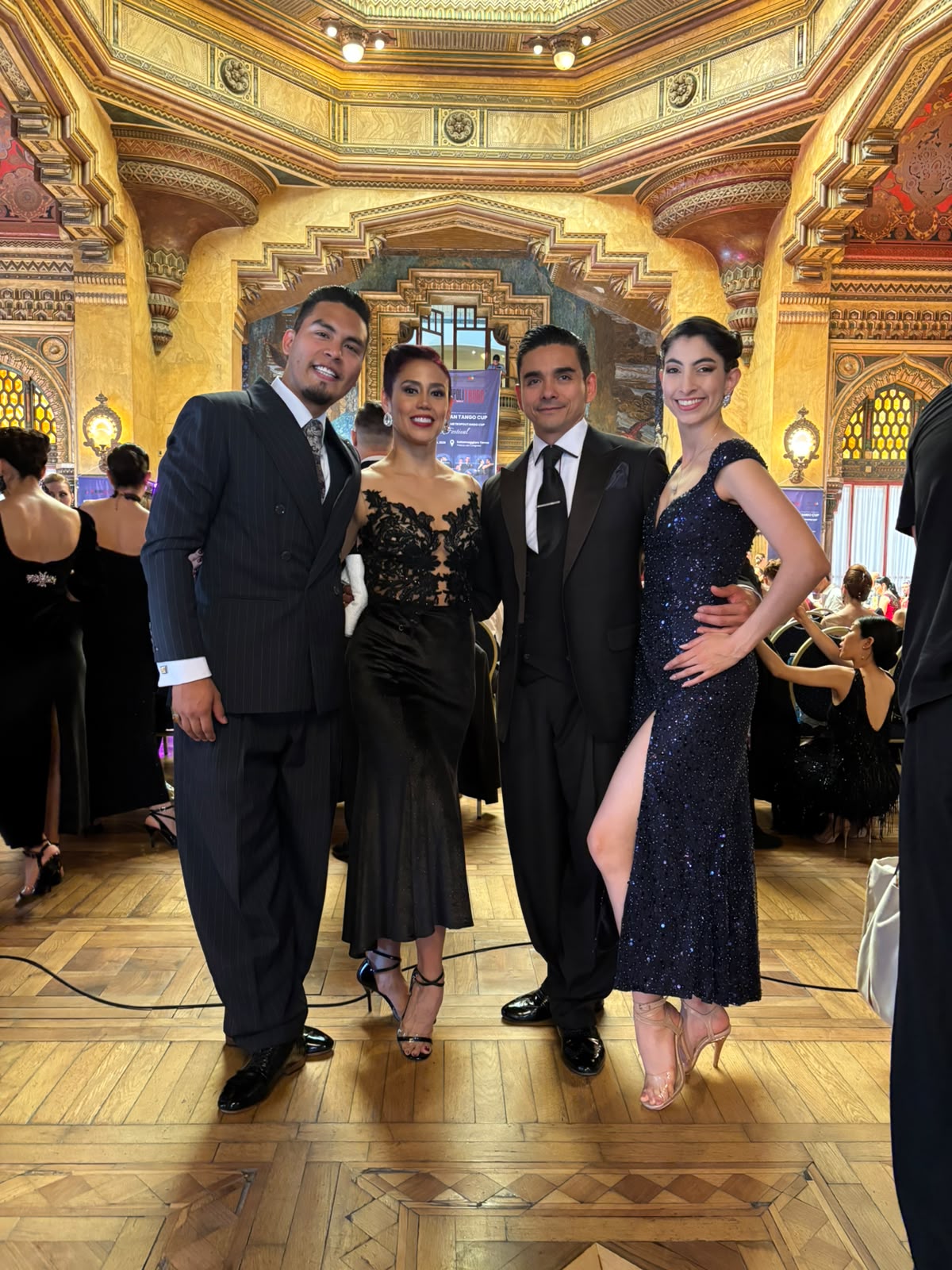 Four adults in formal attire pose for a group photo at a lavish, gilded ballroom. 0