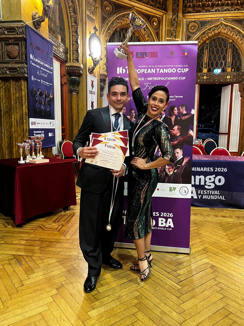Award ceremony: a man in a suit holds certificates and a medal while a woman in a sparkling black dress raises a trophy beside a purple banner and ornate hall backdrop for a tango competition.