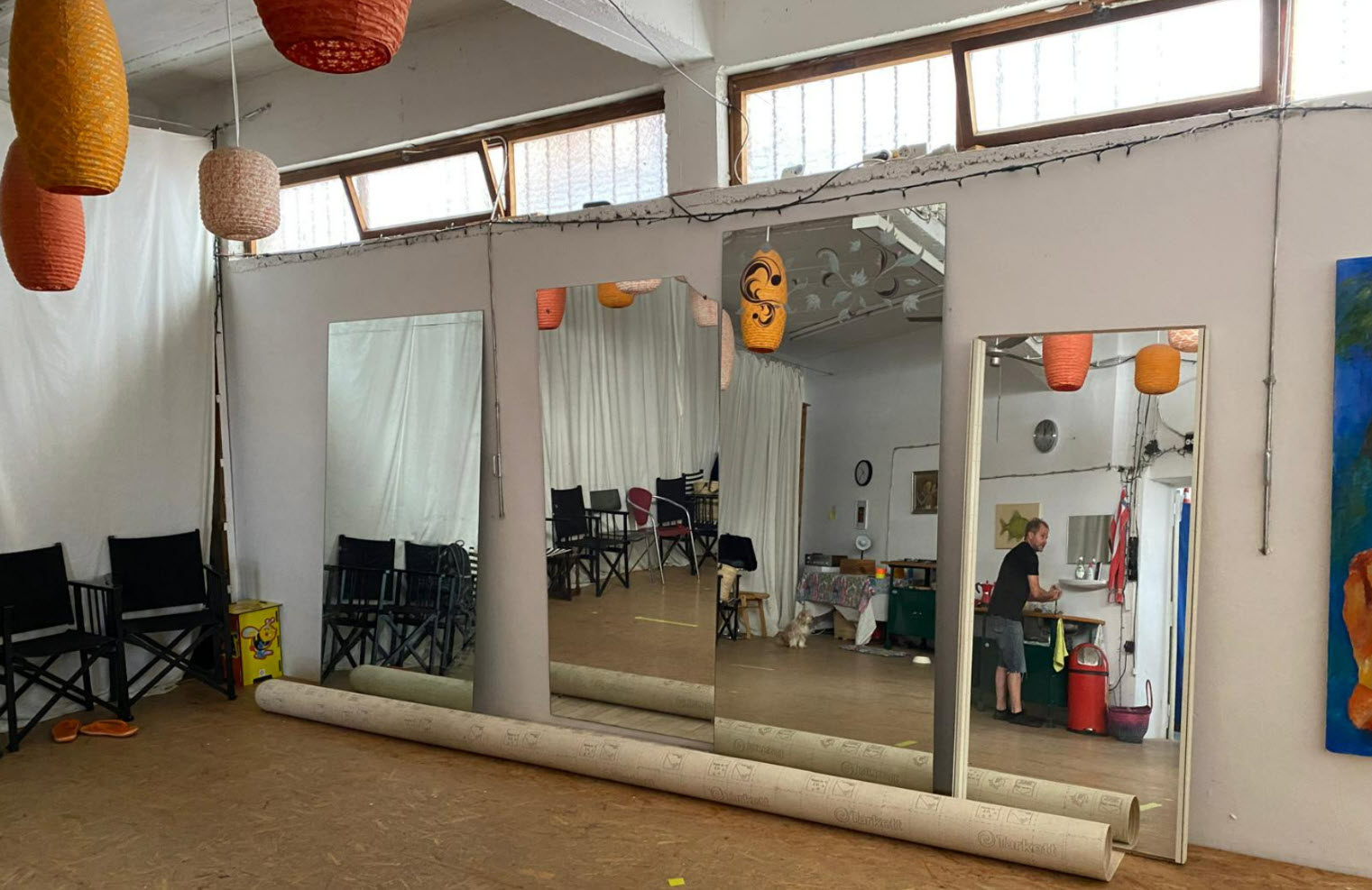Rehearsal studio with large mirrors, stacked folding chairs, and rolled rugs along the floor; orange paper lanterns hang from the ceiling.